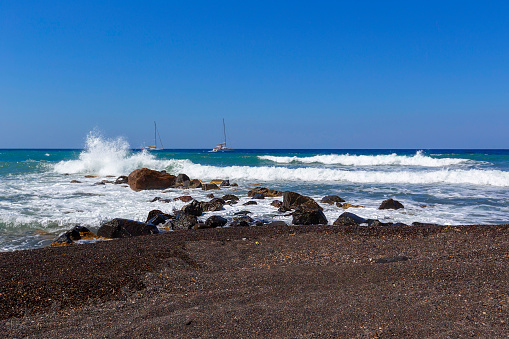 Santorini, beautiful black beach of Vlychada illuminated by the sun. Beautiful sea with high long waves breaking on black lava rocks. In the background two white sailboats.