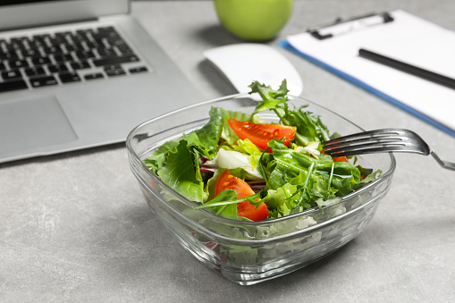 Fresh vegetable salad, laptop and fork on light grey table at workplace, closeup. Business lunch