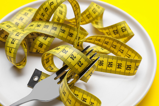 Measuring tape and fork on yellow background, closeup. Weight loss concept