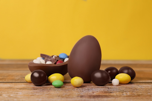 Delicious chocolate eggs and colorful candies on wooden table against yellow background