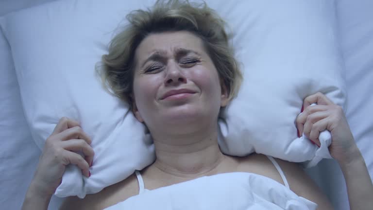 Irritated deeply upset woman adjusting ugly pillow, lying in bed of cheap hotel, has problems with comfort, can't fall asleep because feeling pain in neck and shoulders