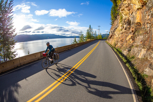 A man goes road bicycle touring on a quiet, mountain road in British Columbia, Canada. He has panniers and a handlebar bag on his bike to carry camping gear.
