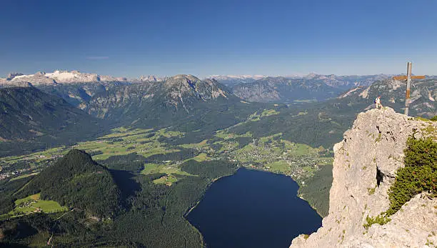 "Panorama from one of the highest mountains in the Austrian Alps. You can see the summit cross on the right with an older woman beside it who has just reached the top. In back you see the glacier dachstein, most of the Alps and the lake Altaussee in the middle of the nature reserve Ausseerland / Salzkammergut."