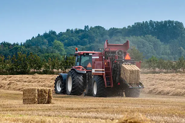 A tractor is pulling a straight-line straw baling machine. A forest can be seen in the background.