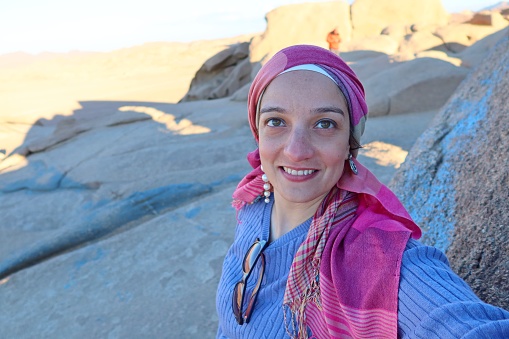 A cheerful young woman wearing a purple pullover and a head cover  with hazel eyes taking a selfie in the middle of the rocks of the blue desert in Egypt