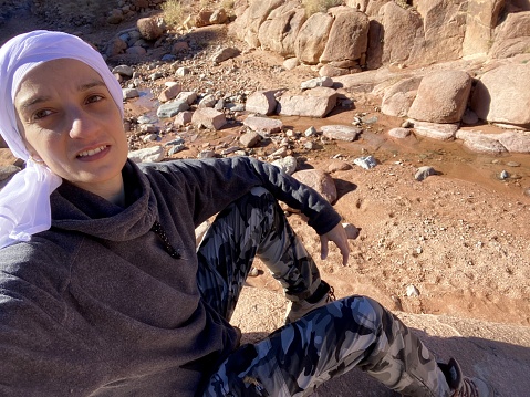 A cheerful young woman wearing grey fleece and a white head piece with hazel eyes taking a selfie while hiking to the top of  saint Catherine mountain  the highest peak  in Egypt