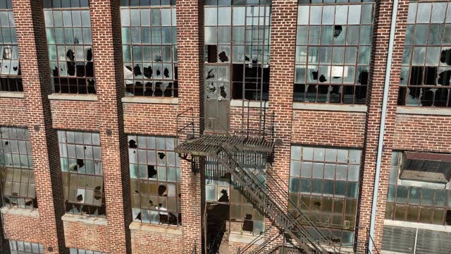 Close up view of old brick industrial building facade with shattered windows and a rustic metal fire escape, conveying urban decay and historical architecture. Aerial establishing shot during sunset.