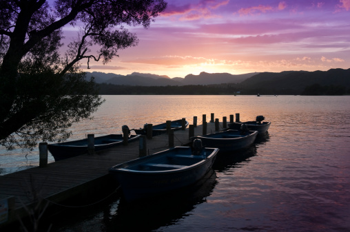 Sun sinking over the cumbrian hills send reflections of a striking sunset acroos the waters of windermere lake. Moored row boats in foreground