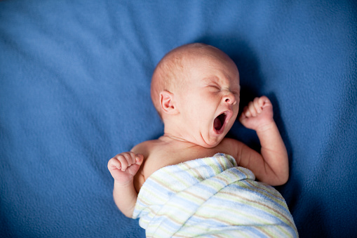 Color photo of a tired newborn baby yawning and stretching in a striped blanket on a soft, blue blanket.