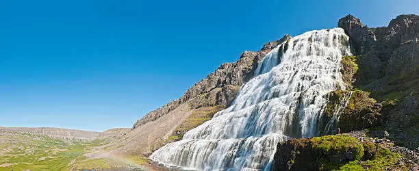"Child in colorful red jacket dwarfed by the massive roaring cascades, bright white torrents and towering cliffs of the Dynjandi (Fjallfoss) falls high in the remote Westfjords of wild Iceland under clear blue panoramic skies. ProPhoto RGB profile for maximum color fidelity and gamut."