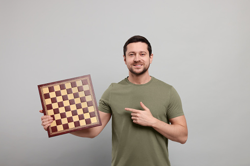 Handsome man pointing at chessboard on light grey background