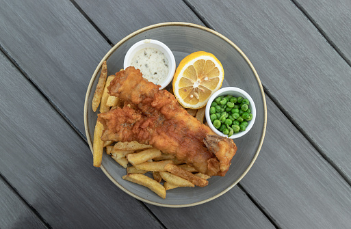 Authentic traditional british cuisine Fish and Chips served with French fries, Green peas, cut Lemon and Tartar sauce on Ceramic plate. Top view, Space for your text, Selective focus.