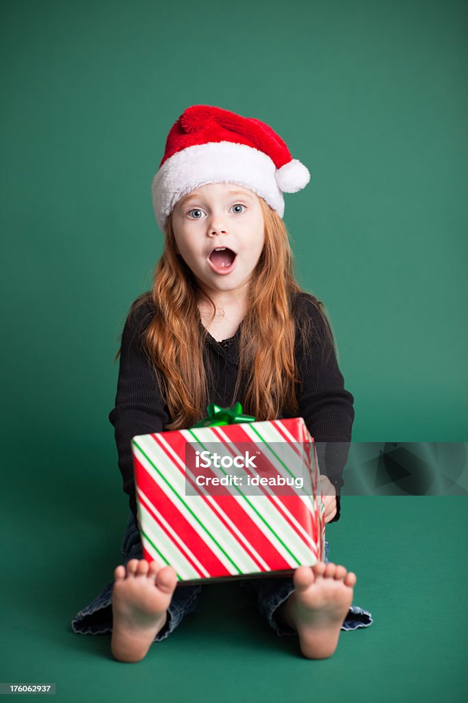 Surprised Girl Wearing Santa Hat with Christmas Present Color image of a surprised 4-year-old girl wearing a Santa hat and sitting with a Christmas present. Isolated on green background. 2-3 Years Stock Photo