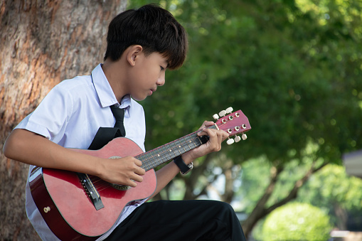 Asian handsome schoolboy is sitting in school park and playing acoustic guitar or ukulele, soft focus, concept for young teenager with popular instruments.