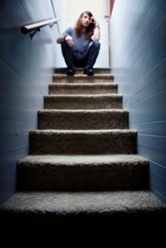 A dark and moody contrast picture of a lonely young adult man sitting at the top of a stairway.  Focus is on the foreground steps.  Vertical with copy space.
