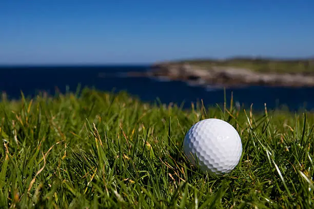 A golf ball lays in longer grass off the fairway in a beautiful ocean setting on a bright blue sky day. Focus on ball with copy space.