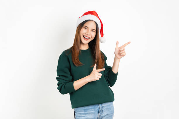 Cheerful beautiful Asian woman wearing red Christmas hat and hand pointing to the side to copy space on white background. stock photo