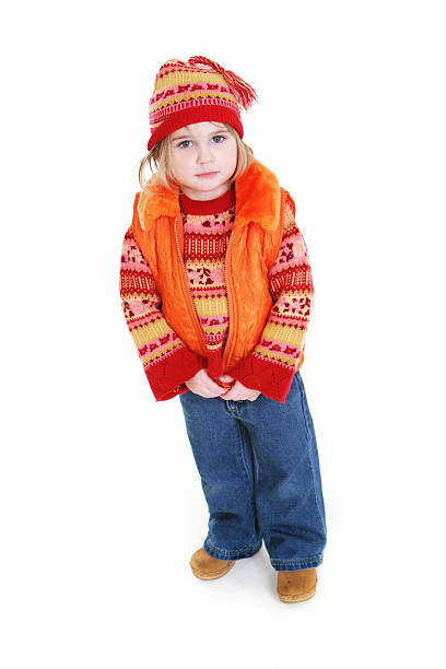 Little girl in cold weather clothing looking at camera from a down angle view. Sweater, vest, cap and jeans. Full body isolated on white.