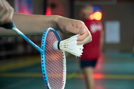 Badminton player holds white shuttlecock in hand, standing and serving it over the net to another side of badminton court.