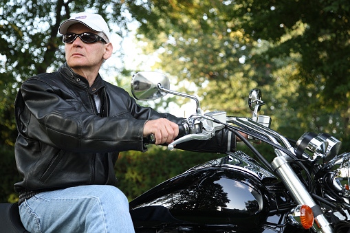 Smiling man biker holding helmet and looking at camera on sport motorcycle. Handsome successful fashion model wearing stylish leather jacket posing for pictures