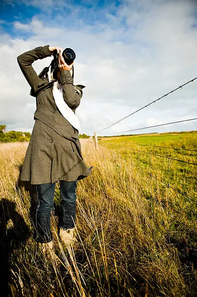Photographer woman composing photo farm fence outdoors. The woman is wearing a himalayan woollen beanie and an overcoat.