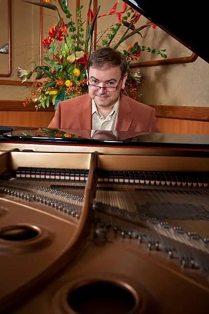 A friendly-looking piano player at a grand piano.  He looks directly at the camera with a quizzical look.