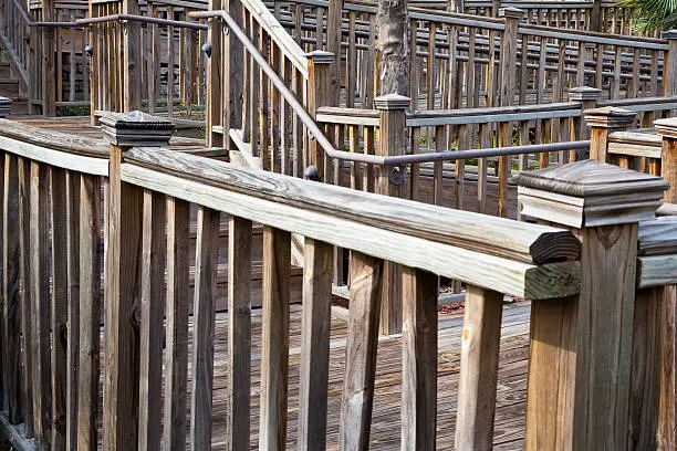 faded wooden guardrails zig zagging up walkway and around a wooden deck. The wood is weathered to gray or light tan in some areas. some balusters are loose and crooked.