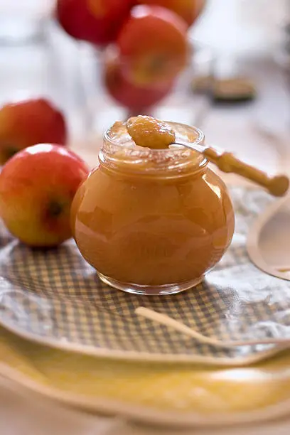 A jar of homemade applesauce with a spoon balanced on the rim.  The jar sits on a stack of colorful gingham bibs and apples are scattered in the background.