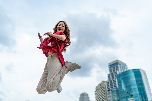 Young Asian woman holding mobile phone and jumping in urban city. Lifestyle outdoor activity concept.