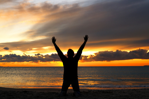 Man with arms raised kneeling on the beach at sunset.