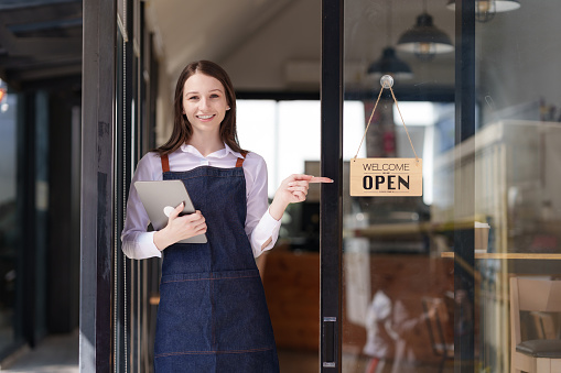 Portrait of a happy waitress standing at restaurant entrance. Happy woman owner showing open sign in her small business shop.