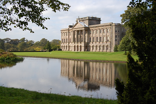 Image of a traditional large English mansion house with portico entrance. In the foreground a lake moat with reflection of the mansion hall. Lyme Park Hall, Cheshire, UK.