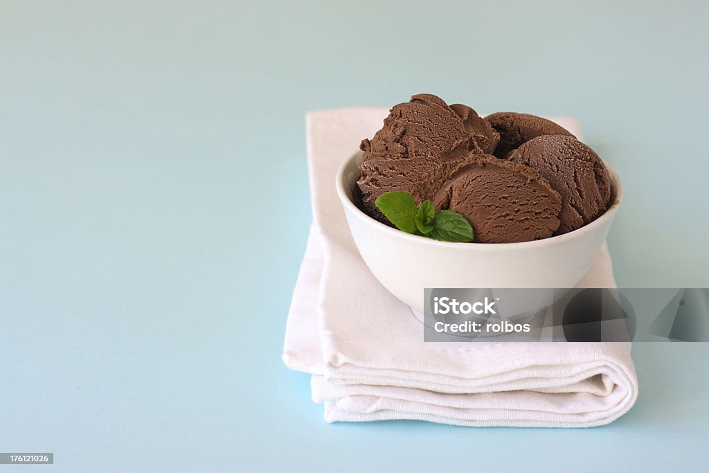 chocolate ice cream in white bowl "Scoops of chocolate ice cream with mint leaves in a white bowl standing on a folded white napkin on light blue background. This is real ice cream so some ice crystals are visible as well as a little bit of smudging on the rim of the bowl. Shallow depth of field, focus on left hand scoop." Bowl Stock Photo