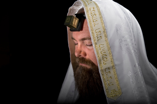Jewish Rabbi Closes His Eyes in Prayer. Wearing Tallit (Prayer Shawl) with a Beautiful Gold Crown. Tefillin with leather strap is worn on the head. Isolated on blackSee Images Below: