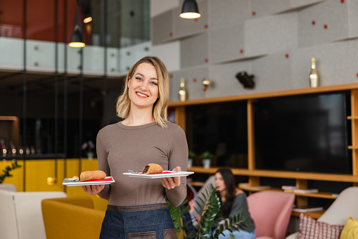 The attractive Caucasian female bartender working in a cosy colourful cafe looking at the camera while holding two plates with desserts on them. She is wearing an apron while working. The woman looks happy and charming.