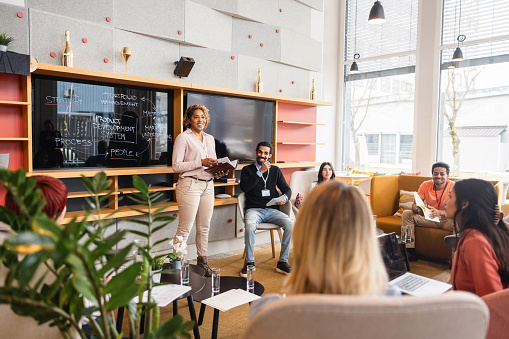 A confident black adult female CEO hosting a weekly business meeting in a cosy co-working space. She is standing in front of the blackboard surrounded by the department managers. Everyone is carefully listening