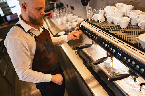 A professional adult male barista using the coffee maker at a cafe to prepare two cups of coffee for his customers. He is wearing a brown apron while working to protect his clothes. Due to the hot temperatures, there is steam as the coffee machine is running.