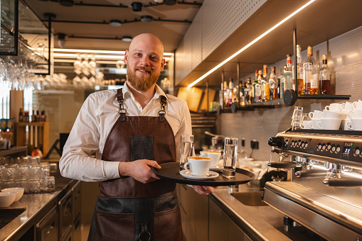 A portrait of a content Caucasian male barista looking at the camera while holding a serving tray with drinks. He is standing behind the counter and is wearing a brown leather apron.