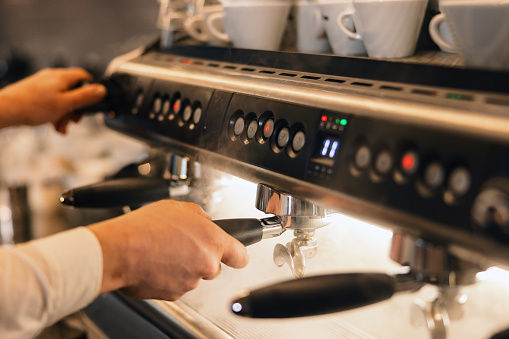 Close up of a Caucasian male barista brewing fresh coffee on a coffee machine in a cafeteria. The coffee machine is expensive and professional. There is a LED light illuminating the working area. The coffee machine is hot and there is steam everywhere thus the barista is careful while preparing coffee.