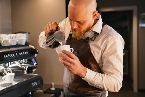 A professional Caucasian adult male barista being careful and precise while creating latte art at a cafeteria. His hands are steady and still while pouring the milk into the coffee. Talented male barista in a small coffee shop.