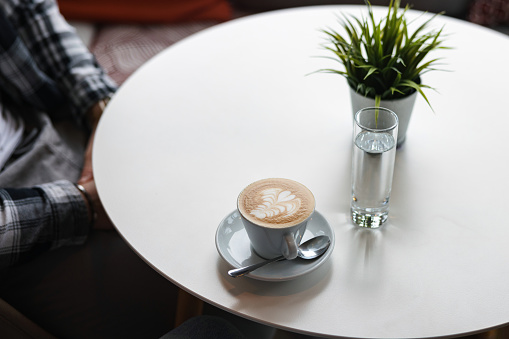 A cup of coffee with latte art at a coffee shop. The coffee cup is white and is placed on a small white plate. There is a silver spoon on it and a glass of water next to the coffee. The table is white.