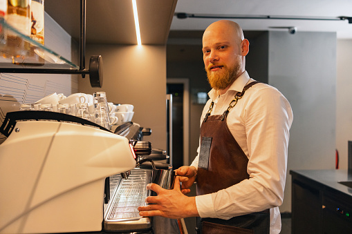 A Caucasian male barista at a cafe looking at the camera while standing by the coffee maker. He is using the coffee maker to froth some milk in order to to make a cappuccino for a customer. The man looks experienced. He is wearing a classy shirt and a brown leather apron.