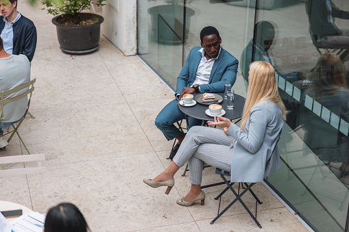 A professional black businessman talking to his Caucasian female colleague while they are sitting at a cafe. They are located outdoors. The business people are enjoying their delicious coffee while having a serious conversation about the company.