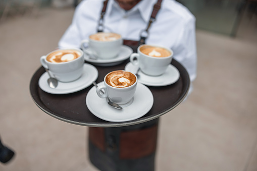 A black tray with four cups of coffee held by a male waiter in an apron. The coffee cups are white. There is latte art on the coffees