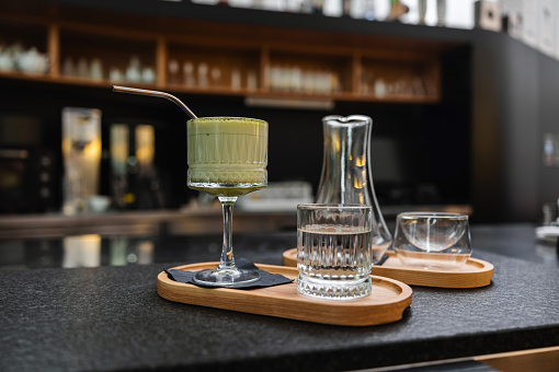 Beautiful glass of matcha drink with a straw and a serving of filtered coffee on a fancy wooden tray. The trays are placed on a counter of the modern cafeteria.
