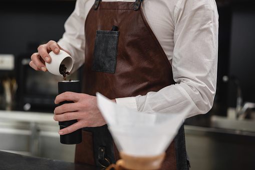 Close up of a Caucasian male barista pouring coffee beans into a coffee grinder before making a coffee for a customer. The face of the barista is not visible. He is wearing a brown leather apron and a white shirt.
