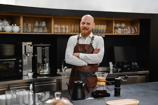 A professional Caucasian male barista standing by the counter with his arms crossed and looking at the camera. He is wearing a brown leather apron. He is located at a charming specialty coffee shop and behind the bartender there are glasses stacked on the shelves. The barista looks relaxed and content.