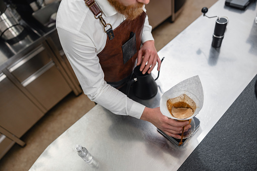 An adult caucasian male barista at a specialty coffee shop preparing coffee with a pour-over technique. He is wearing an apron while working at the cafetaria.