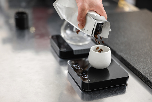A close-up of a scale and a white ceramic cup at a specialty coffee shop. Roasted coffee beans about to be weighed by a barista before making a coffee.