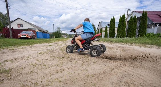 A child makes a sharp turn on an ATV, kicking up dust and sand from the road. The wheels of the ATV slip on the sand, raising dust and sand. A child boy enjoys riding an ATV..